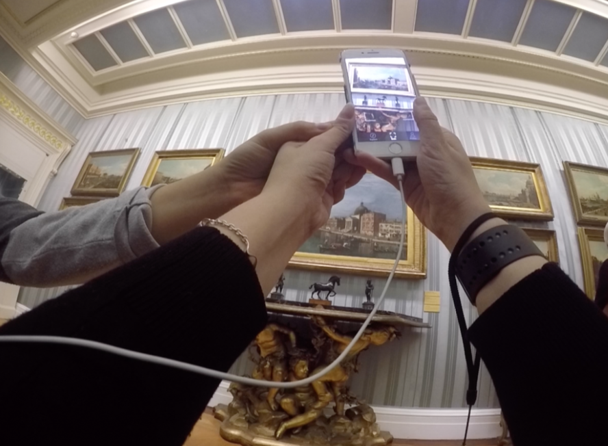 A person uses an AI smartphone app to scan a framed painting in an art gallery, while another person’s hand reaches in to help guide the phone.