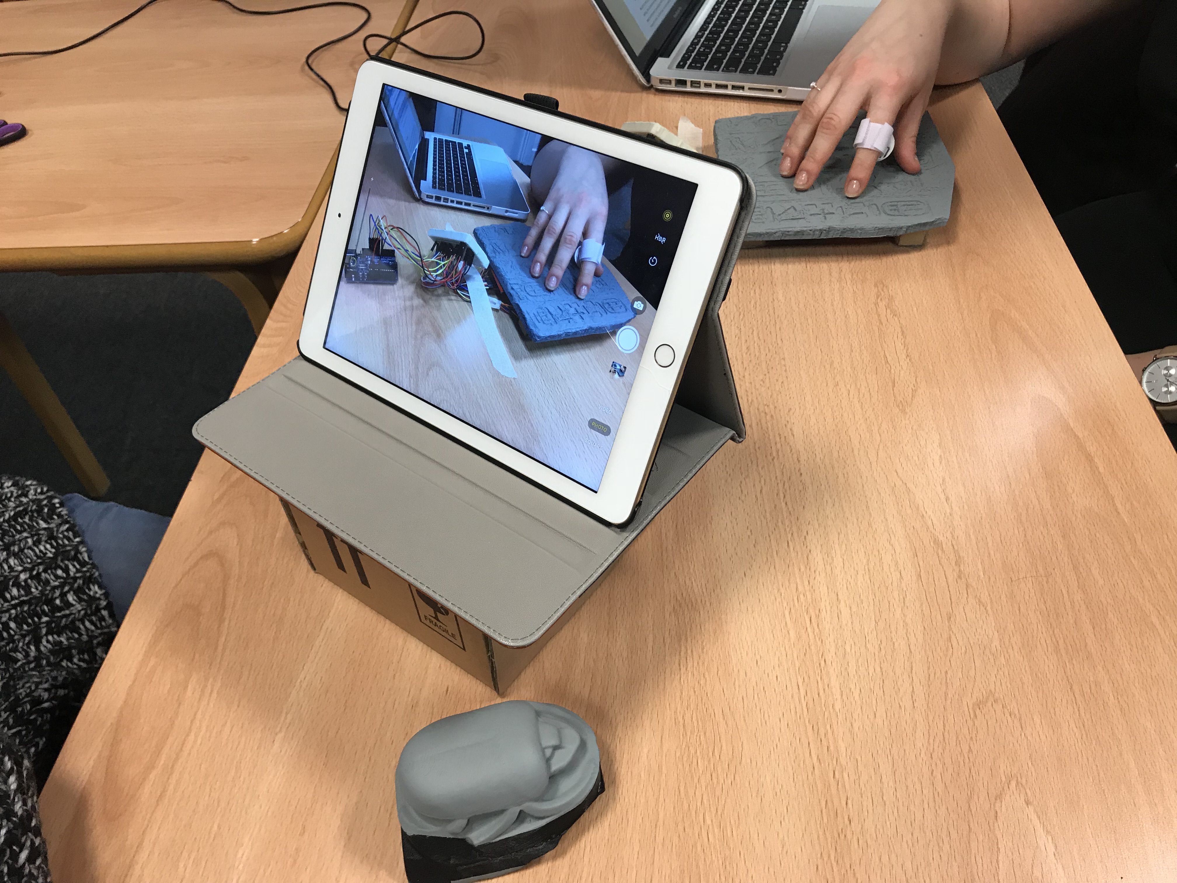 A tablet on a stand shows a live camera view of a person’s hand touching a 3D-printed model of an Ancient Egyptian stele, with a sensor attached to one finger and wires connected to a small electronic board nearby. The real hand mirrors the view on screen as part of a hands-on demonstration. In the foreground, a separate grey 3D-printed model of an Ancient Egyptian scarab beetle sits on the table, with a laptop and other materials in the background.
