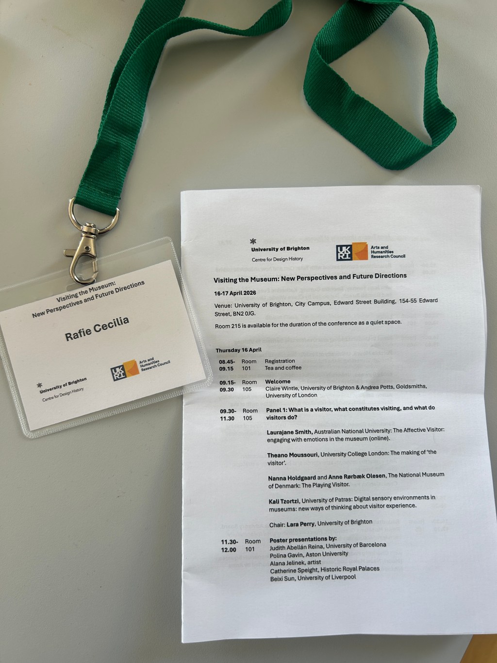 A green lanyard with a conference name badge lies next to a printed conference programme on a white table. The badge reads “Rafie Cecilia” and “Visiting the Museum: New Perspectives and Future Directions.” The program, from the University of Brighton and UKRI Arts and Humanities Research Council, shows the conference title, dates (16–17 April 2026), venue details, and a schedule of sessions.