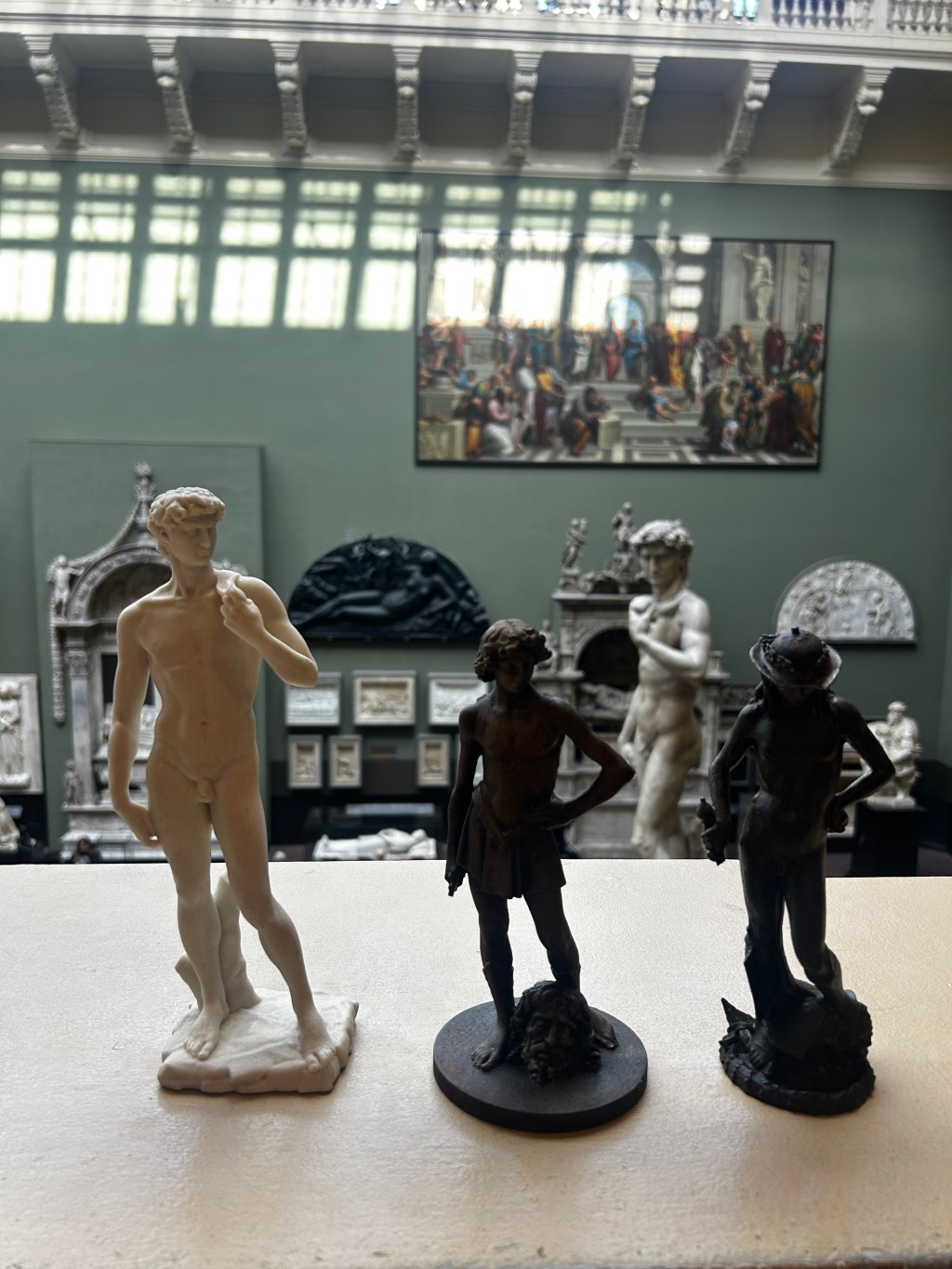 **Alt text:** Three 3D-printed replicas of Renaissance sculptures of David are displayed on a light-coloured table in the foreground of the V&A Museum Cast Courts. The largest replica, a pale-coloured model of Michelangelo’s *David*, stands on the left. Beside it are darker replicas of Donatello’s and Verrocchio’s *David*, each shown standing above the severed head of Goliath. In the background, the Cast Courts are visible with large plaster casts, architectural fragments, and a monumental cast of Michelangelo’s *David*. High windows cast soft light across the gallery, and Raphael’s *School of Athens* tapestry hangs on the far wall. The photograph highlights the contrast in scale between the handheld replicas and the monumental casts displayed within the gallery.
